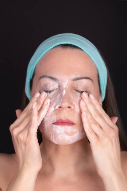 Makyaj merhemiyle yüzünü silen bir kadın, gözleri kapalı ve mavi saç bandı takıyor. Siyah arka plan. Yüz bakımı ve temizlik rutini kavramı