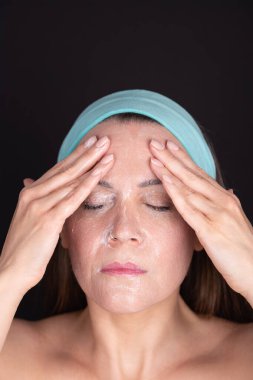Mavi bantlı bir kadının portresi, elleriyle yüz kremi sürerek makyajını temizliyor. Kapalı gözler ve rahat bir ifade. Siyah arka plan. Yüz bakımı ve temizlik rutini kavramı