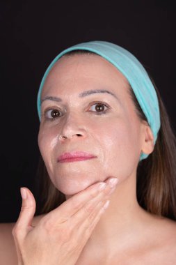 Makyaj çıkarıcı merhem sürerek yüzünü temizlerken kameraya bakan 40 yaşında bir kadın. Saçları mavi saç bandıyla bağlanmış. Siyah arkaplan