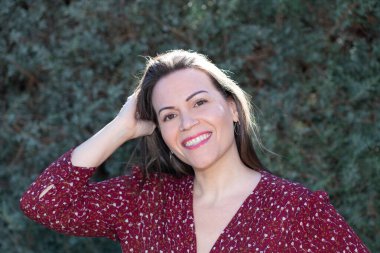 Kırklı yaşlarda, düz kahverengi saçlı bir kadının portresi, kameraya bakıp saçlarına dokunurken gülüyor, neşeli ve tasasız bir ifadeyle. Yeşil bitki örtüsü ve yumuşak doğal ışık.