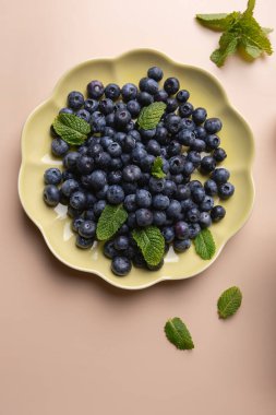 Taze yaban mersinli tabak ve bej arka planda nane yaprakları. Zarif ve canlı meyve kompozisyonu. Sağlıklı beslenme ve tazelik kavramı