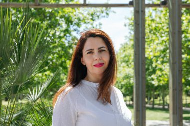 Günlük kıyafetlerle gülümseyen kadın, yeşil bir alanda poz veriyor ve arka planda tahta bir yapı var. Uzayı kopyala Refah kavramı, pozitif enerji ve doğayla temas.