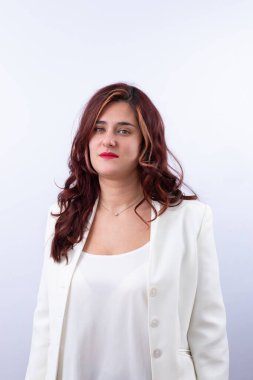 Beyaz takım elbiseli, kırmızı dudaklı, uzun dalgalı kumral saçlı ve kameraya özgüvenle bakan zarif bir kadının portresi. Tarafsız arka plan, profesyonel ve modern tarz. İş için ideal, kadın gücü ve moda. Boşluğu kopyala. 