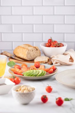 Bir tabak avokado ve kiraz domatesli mermer masa. Arkaplanda tam tahıllı ekmek, domates kasesi ve yumuşak bez var. Taze ve dengeli kompozisyon. Sağlıklı gıda ve doğal yaşam tarzı kavramı