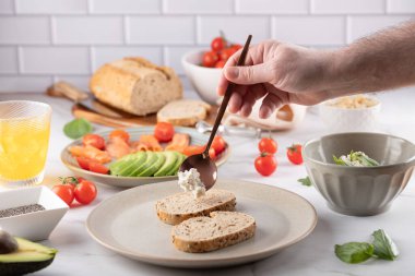 Erkek el, kepekli ekmeğin üzerine krem peynir koyuyor. Arka planda taze malzemeler ve bir bardak turuncu Kombucha var. Ev yemeği ve sağlıklı yaşam tarzı.