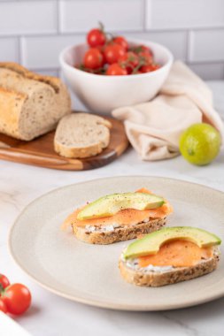 Krem peynirli tam tahıllı tost, somon ve avokado bej bir tabakta, ekmek, vişneli domates ve arka planda limon ile servis edilir. Minimalist tarzda sağlıklı bir kahvaltı.