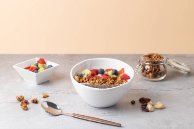 Kefir, granola, chia tohumu ve taze böğürtlenlerle dolu beyaz seramik kâse, yanında da küçük kare bir kase dolusu meyve ve taş bir yüzey üzerinde granola bulunan cam bir kavanoz. Sıcak bej renkli arka plan. Sağlıklı kahvaltı kavramı 