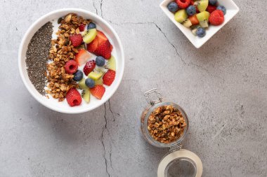 Kahvaltının beyaz kefir, meyve, granola ve chia tohumları, ahşap ve metal kaşık, tahıl gevrekli kavanoz ve hafif taşlı arka planda meyveli kare kâse manzarası. Sağlık ve sağlıklı yaşam kavramı