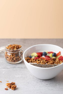 Hafif taş yüzeyinde kefir, chia tohumu, granola ve meyveli sağlıklı kahvaltı kasesi. Küçük bir kavanoz granola ve biraz dağınık granola parçaları olay yerine doku katıyor. Sağlıklı kahvaltı ve modern yaşam tarzı kavramı