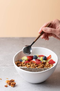 Kefir, çilek, kivi, ahududu, böğürtlen, yaban mersini, granola ve chia ile beyaz kâse üzerinde kaşık ile kadın eli. Şeftali arkaplan ve gri taş yüzey. Sağlıklı beslenme kavramı