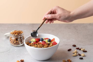 Beyaz kefir, çilek, böğürtlen, ahududu, kivi, granola ve chia tohumlarıyla sağlıklı bir kahvaltı. Gri taş yüzeyde kaşık tutan kadın eli. Modern yaşam tarzı ve dengeli beslenme kavramı