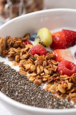 Beyaz kasede besleyici kahvaltı ayrıntıları: kefir, dilimlenmiş meyve, altın granola ve chia tohumları. Arkaplanı granola kavanozuyla bulanık. Dikkatli yeme ve sağlıklı yaşam tarzı kavramı