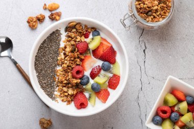 Kefir, granola, chia ve taze meyveli beyaz kâse manzarası. Solda bir metal kaşık, sağda bir açık granola kavanozu ve bir kase meyve. Doğal kahvaltı ve işlenmemiş gıda kavramı