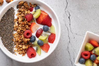 Gri taş yüzeyinde kefir, granola, chia tohumu ve karışık meyveli beyaz kâse manzarası. Sağda, daha fazla meyveli küçük bir kase. Sağlıklı kahvaltı ve dengeli yemek kavramı