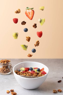 Beyaz kasede çilek, ahududu, kivi ve yabanmersini, kefir, chia tohumu ve granola gibi meyveler havada asılı. Şeftali rengi arka plan ve taş masa. Sağlıklı kahvaltı ve enerji kavramı 