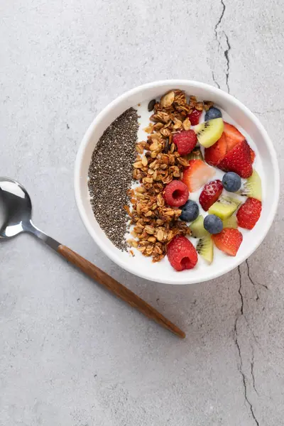 Granola, chia tohumu, çilek, ahududu, kivi ve yaban mersinli kefir kasesinin üst görüntüsü. Gri taş zemin üzerinde ahşap ve metal kaşık var. Dikey görüntü. Sağlıklı beslenme ve tazelik kavramı