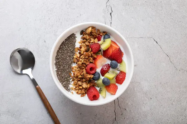 Kefir, çilek, kivi, ahududu, yaban mersini, chia tohumu ve granola içeren beyaz kâse, gri taş zemin üzerinde metal ve ahşap kaşık. Sağlıklı kahvaltı ve modern yaşam tarzı kavramı