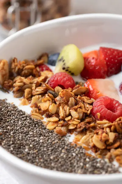 Beyaz kasede besleyici kahvaltı ayrıntıları: kefir, dilimlenmiş meyve, altın granola ve chia tohumları. Arkaplanı granola kavanozuyla bulanık. Dikkatli yeme ve sağlıklı yaşam tarzı kavramı
