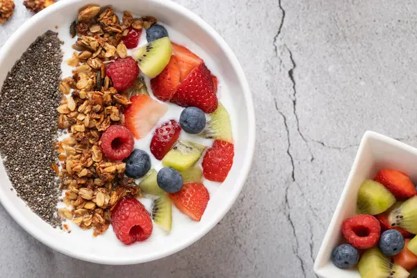 Gri taş yüzeyinde kefir, granola, chia tohumu ve karışık meyveli beyaz kâse manzarası. Sağda, daha fazla meyveli küçük bir kase. Sağlıklı kahvaltı ve dengeli yemek kavramı