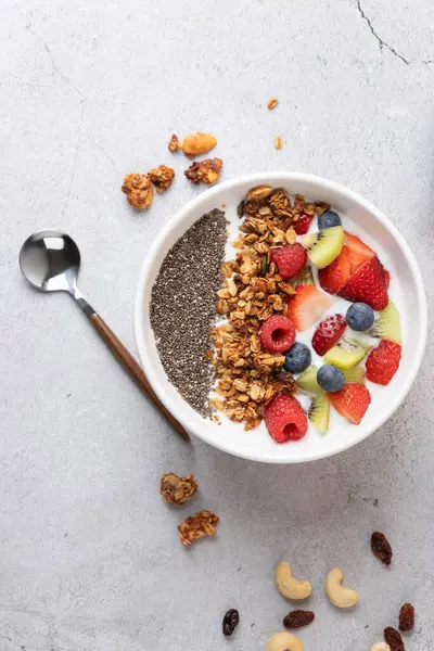 Gri yüzeyinde kefir, taze meyve, granola ve chia tohumları olan beyaz bir kasenin dikey aşağı çekimi. Solda, metal bir kaşık; altta, fındıklar. Sağlıklı kahvaltı ve gerçek yemek kavramı
