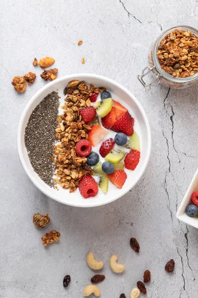 Gri taş zemin üzerinde kefir, meyve, chia ve granola olan bir kasenin dikey üst görüntüsü. Dağınık fındıklar ve iki görünür konteynırla dikkatlice düzenlenmiş kompozisyon. Sağlıklı beslenme ve doğal yaşam tarzı kavramı