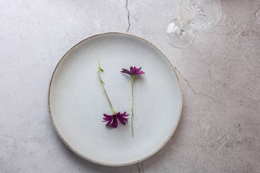 Açık gri seramik tabağın üst görüntüsü. İki mor çiçek ve boş bir kristal bardak. Uzayı kopyala Zarif masa düzenleme ve çiçek minimalizmi kavramı