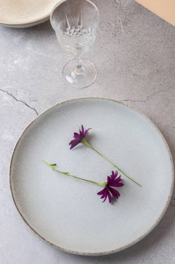 Gri taş yüzeyinde iki menekşe çiçekli seramik tabağın üst görüntüsü. Sahnede kristal cam var. Minimalist masa kompozisyonu. Doğal zarafet ve sükunet kavramı