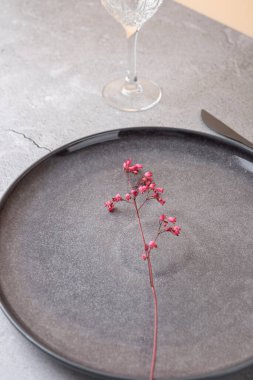 Koyu gri bir seramik tabağın yakın görüntüsünde küçük pembe bir çiçek var. Kesik bir kristal cam arka planda kısmen görülebilir. Hafif beton yüzeyde basit bir bileşim. Hassas masa üstü ayrıntısı ve minimum gıda tasarımı kavramı