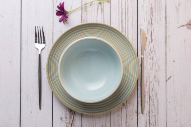 Açık mavi seramik bir tabağın yatay görüntüsü çatal bıçak takımı ve açık ahşap bir zeminde mor bir çiçek. Uzayı kopyala Zarif masa düzeni, minimalizm ve İskandinav tarzı.