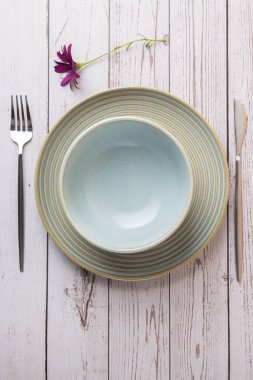 Açık mavi bir seramik tabağın üst görüntüsü yemek takımları ve açık ahşap arka planda mor bir çiçek. Zarif masa düzeni, minimalizm, doğal tasarım ve İskandinav tarzı.
