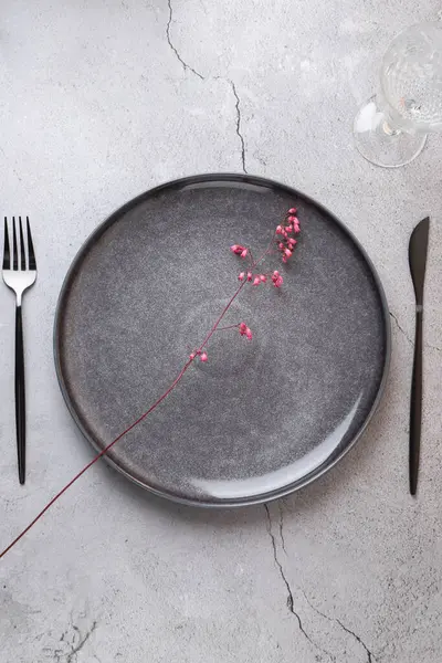 Zarif ve minimalist bir kompozisyonu olan bir masanın yukarıdan görünüşü. Üzerinde modern çatal bıçak takımı ve beton bir yüzey üzerinde cam bardak bulunan biçimlendirilmiş pembe çiçekli gri bir seramik tabak. Uzayı kopyala Rafine masa estetiği kavramı 