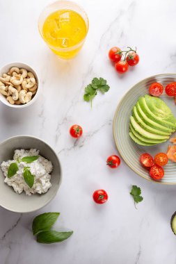 Avokado, kiraz domatesleri, ricotta, kaju ve mermer zemin üzerinde buzlu kombu çayı ile taze ve sağlıklı bir kahvaltı manzarası. Sağlıklı gıda ve minimalist estetik kavramı