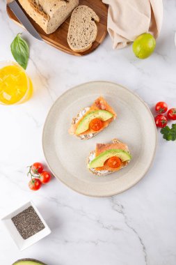 Avokado, somon ve bej tabakta kiraz domatesli tost. Açık mermer arka planda yeni malzemelerle üst manzara. Sağlıklı kahvaltı ve dengeli yaşam tarzı kavramı