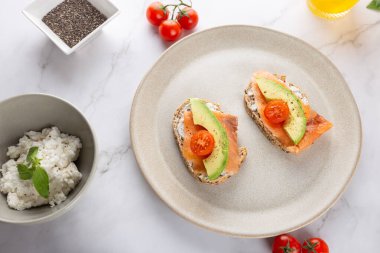 Avokado, somon ve kiraz domatesli tam tahıllı tost, taze malzemelerle beyaz mermer arka planda. Üst Manzara. Sağlıklı kahvaltı ve doğal yaşam tarzı kavramı