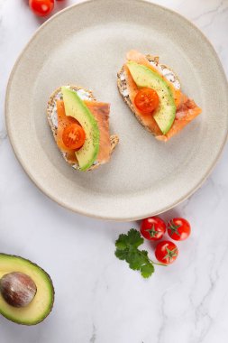 Avokado, somon ve bej tabakta kiraz domatesli iki tostun üst görüntüsü. Beyaz mermer arka plan. Sağlıklı kahvaltı ve dengeli beslenme kavramı