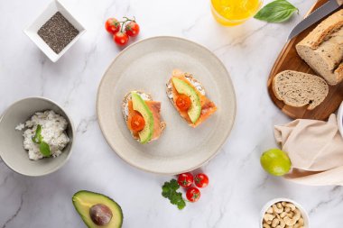 Avokado, somon ve kiraz domatesli tam tahıllı tostların beyaz mermer üzerine taze malzeme ve bir bardak kombu çayı. Sağlıklı gıda ve minimalist estetik kavramı