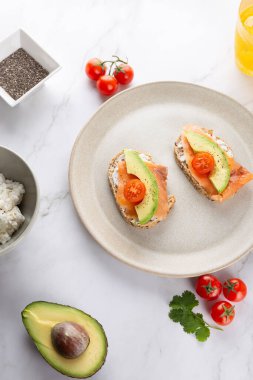 Avokado, somon ve kiraz domatesli tostları bej seramik tabakta mermer yüzeyde malzemelerle birlikte. Uzayı kopyala Sağlıklı kahvaltı ve doğal yaşam tarzı kavramı