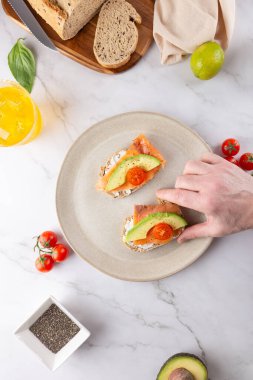 Avokado ve somonla kadeh kaldıran bir erkekle sağlıklı bir kahvaltı. Beyaz mermer üzerine yerleştirilmiş doğal malzemeler. Uzayı kopyala Ev yapımı sağlıklı yiyecek ve aktif yaşam tarzı kavramı