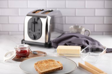 Seramik tabakta tam tahıllı tost, tereyağı ve beyaz mermer tezgahta reçel. Arka planda tost makinesi ve gri bez var. Uzayı kopyala Ev yapımı kahvaltı kavramı