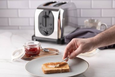 Erkek el, tereyağını bir dilim tostun üzerine seramik tabağa sürüyor. Sabah mutfak sahnesini çilek reçeli, tost makinesi ve gri peçete tamamlıyor. Uzayı kopyala Evde dengeli ve doğal kahvaltı kavramı