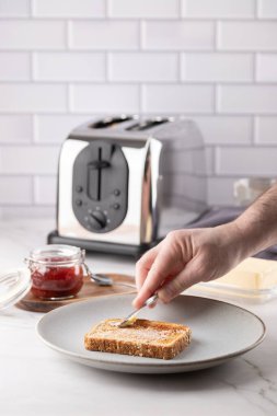 Elle tereyağlı tost dilimi, tereyağı tabağı, çilek reçeli ve arka planda metalik tost makinesiyle. Parlak mutfak tezgahı sahnesi. Uzayı kopyala Ev yapımı kahvaltı ve sabah rutini kavramı