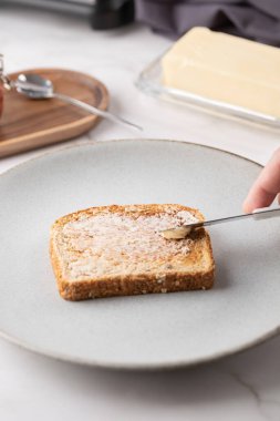 Bir adamın elinin tost ekmeğine tereyağı sürerken yakın plan fotoğrafı, mutfak aletleri ve mermer tezgahta tereyağı tabağı. Kahvaltı hazırlığı ve sağlıklı yaşam tarzı kavramı