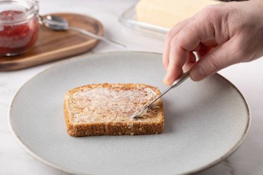 Adamın eli tereyağlı ekmeği gri bir tabağa sürüyor. Tahta, reçel kavanozu ve tereyağı da sahnenin içinde. Doğal kahvaltı ve sağlıklı yaşam tarzı kavramı