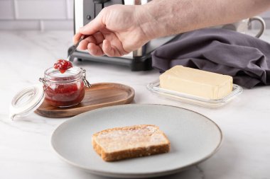 Bir adamın eli, tereyağlı bir tost diliminin önünde çilek reçelli bir kaşığı gri bir tabakta, bir mutfak ortamında mermer bir tezgahın üzerinde tutuyor. Dikkatli yemek kavramı