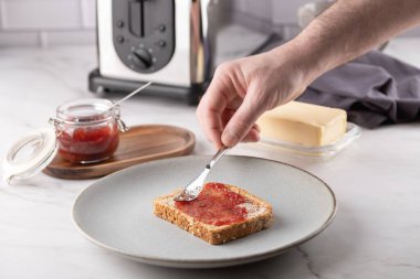 Bir adamın elinin ön görüntüsü, parlak bir mutfakta ekmek kızartma makinesi ve tereyağlı bir bıçakla çilek reçelini ekmeğe yayıyor. Ev yapımı ve sağlıklı kahvaltı kavramı