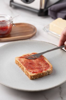 Bir adamın eli, kahvaltılık malzemelerle beyaz mermerin üzerine çilek reçeli serpiştirirken yakın plan. Günlük yemek pişirme ve sağlıklı yaşam tarzı kavramı