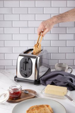 Krom tost makinesinden sıcak tost almak için tahta maşa kullanan bir erkeğin ön görüntüsü. Tereyağı, çilek reçeli, bardak ve gri peçeteyle mermer tezgahın üzerinde. Uzayı kopyala İşlevsel mutfak ve günlük yaşam kavramı