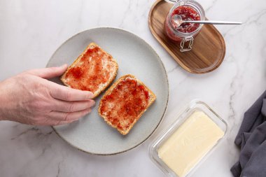 Erkek elinin tost için uzandığı yer gri bir tabakta reçel ve mermer bir yüzey. Reçel kavanozu, tereyağı kabı ve kumaş sahneyi tamamlıyor. Sağlıklı alışkanlıklar ve ev yapımı mutfak kavramı