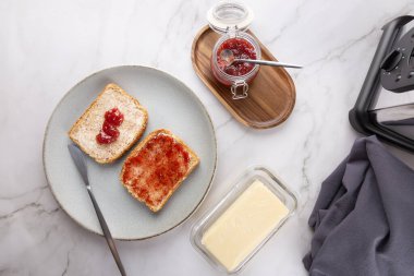 Gri tabakta tereyağı ve çilek reçeli, bıçak, tereyağı, reçel kavanozu, tost makinesi ve kısmen görülebilir gri peçeteli iki tostun üst görüntüsü. Sağlıklı kahvaltı ve günlük mutfak kavramı