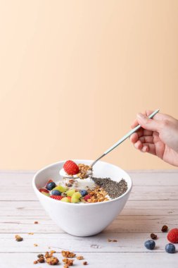 Bir kadının elinde kefir, granola ve içinde taze meyve ve chia tohumları olan bir kaseden alınmış ahududu olan bir kaşıkla dikey görüntüsü. Pastel arkaplan ve kırsal masa. Uzayı kopyala Besleyici kahvaltı ve yaşam tarzı kavramı